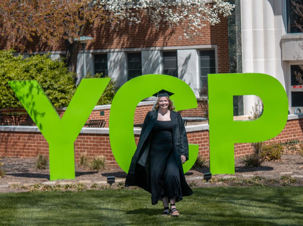 Khora Pawelczyk ’26 stands in graduation attire in front of the large green YCP letters on campus.