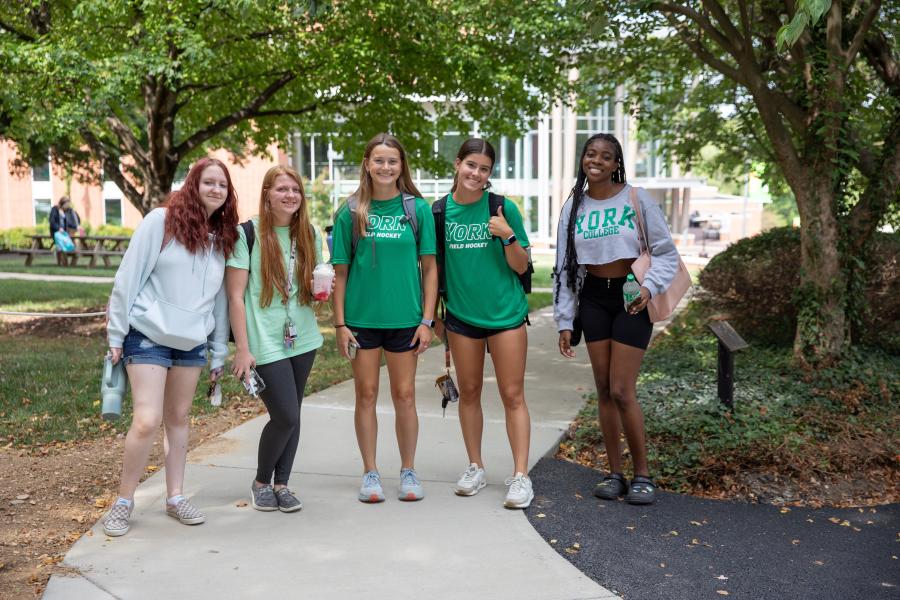 A group of students pose with leafy trees and walkways in the background on a sunny day.