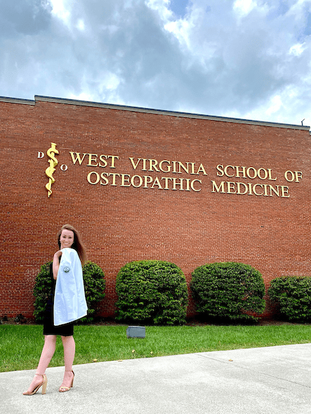 Andrea Smiley poses with a lab coat over her shoulder, standing outdoors beside a sign for West Virginia School of Osteopathic Medicine.