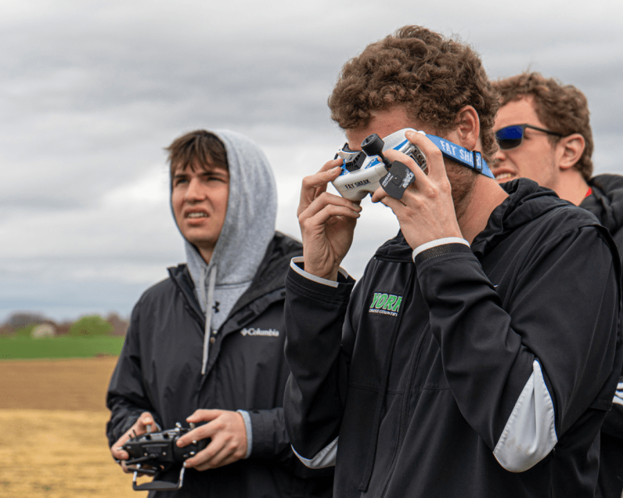 Three people standing outside on a cloudy day, one holding a drone headset to their eyes and another holding a controller.