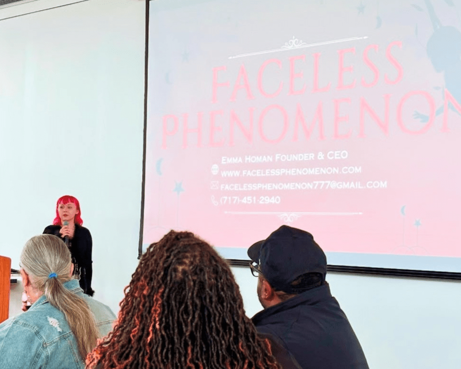 An audience watches a business presentation being given by a person with pink hair, standing in front of a projection screen with the words "faceless phenomenon."