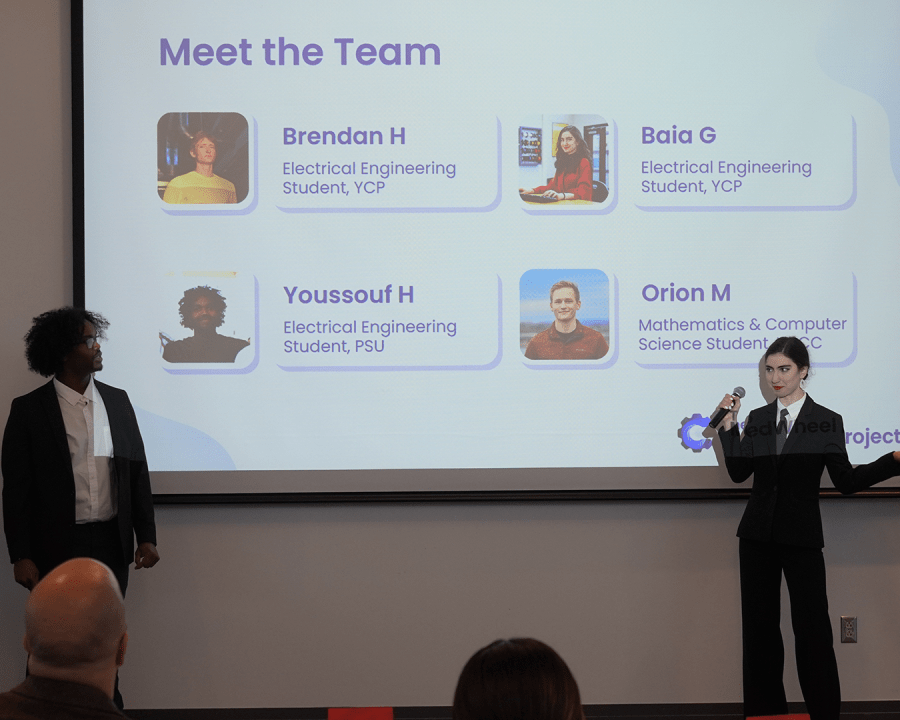 Two people in business attire giving a presentation in front of a projector screen with "Meet the Team" written at the top and introductions below it.