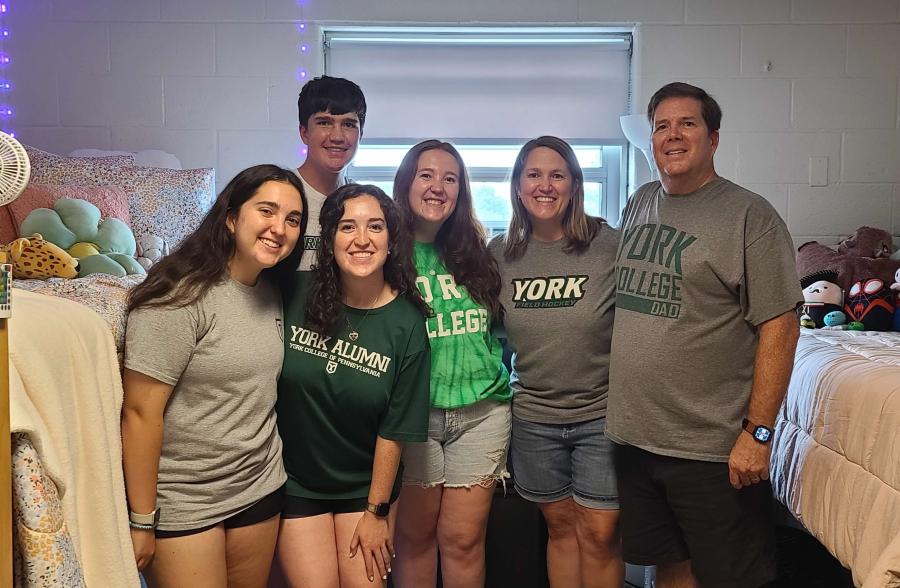 Family wearing York College shirts smile inside a college dorm room.