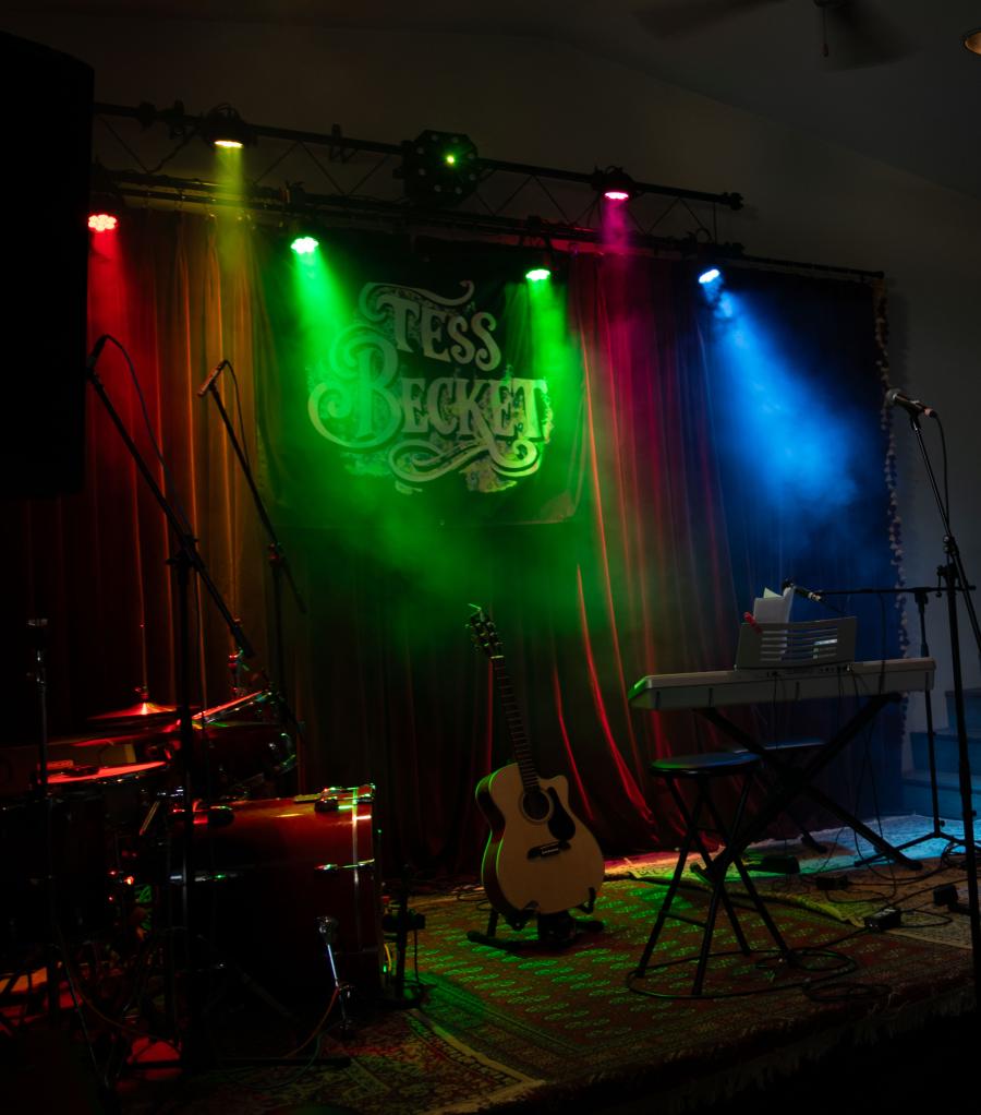 Instruments on a dimly lit stage with colorful lighting and a banner reading "Tess Becket."