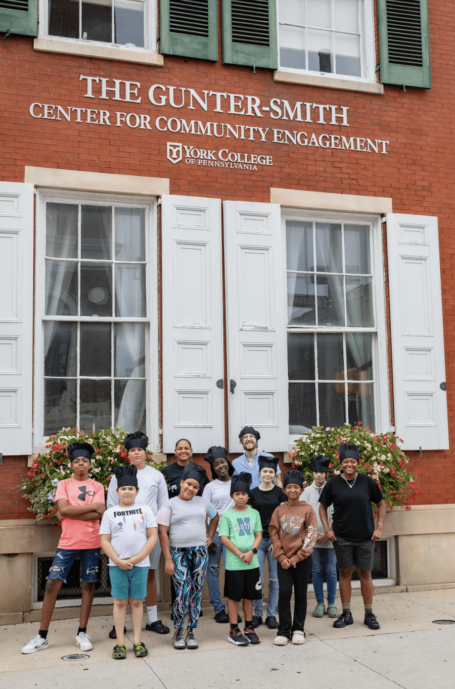 A group of young cooking students in their chef hats stand outside the CCE building.