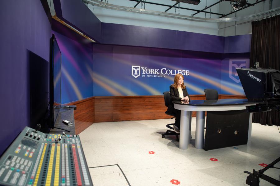 Student in professional attire sitting behind a desk in a TV studio, engaging with cameras.