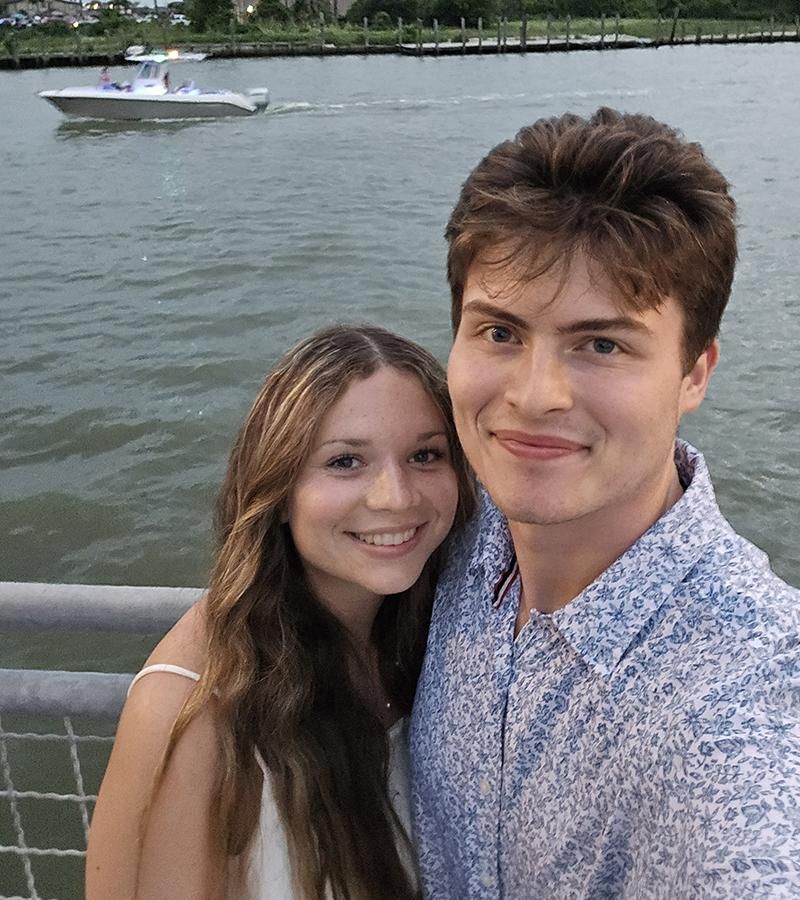 Josh Polancheck stand with his girlfriend with water and a boat behind them