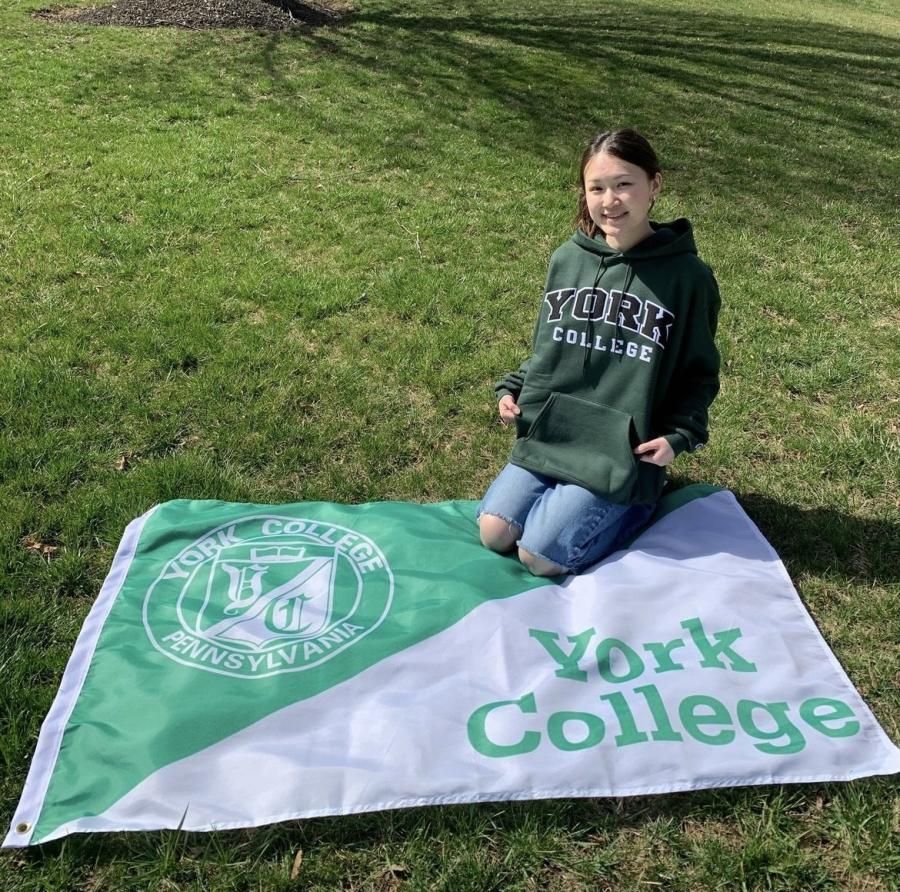 York College student in a green York College hoodie posing with a York College flag in a grassy field.