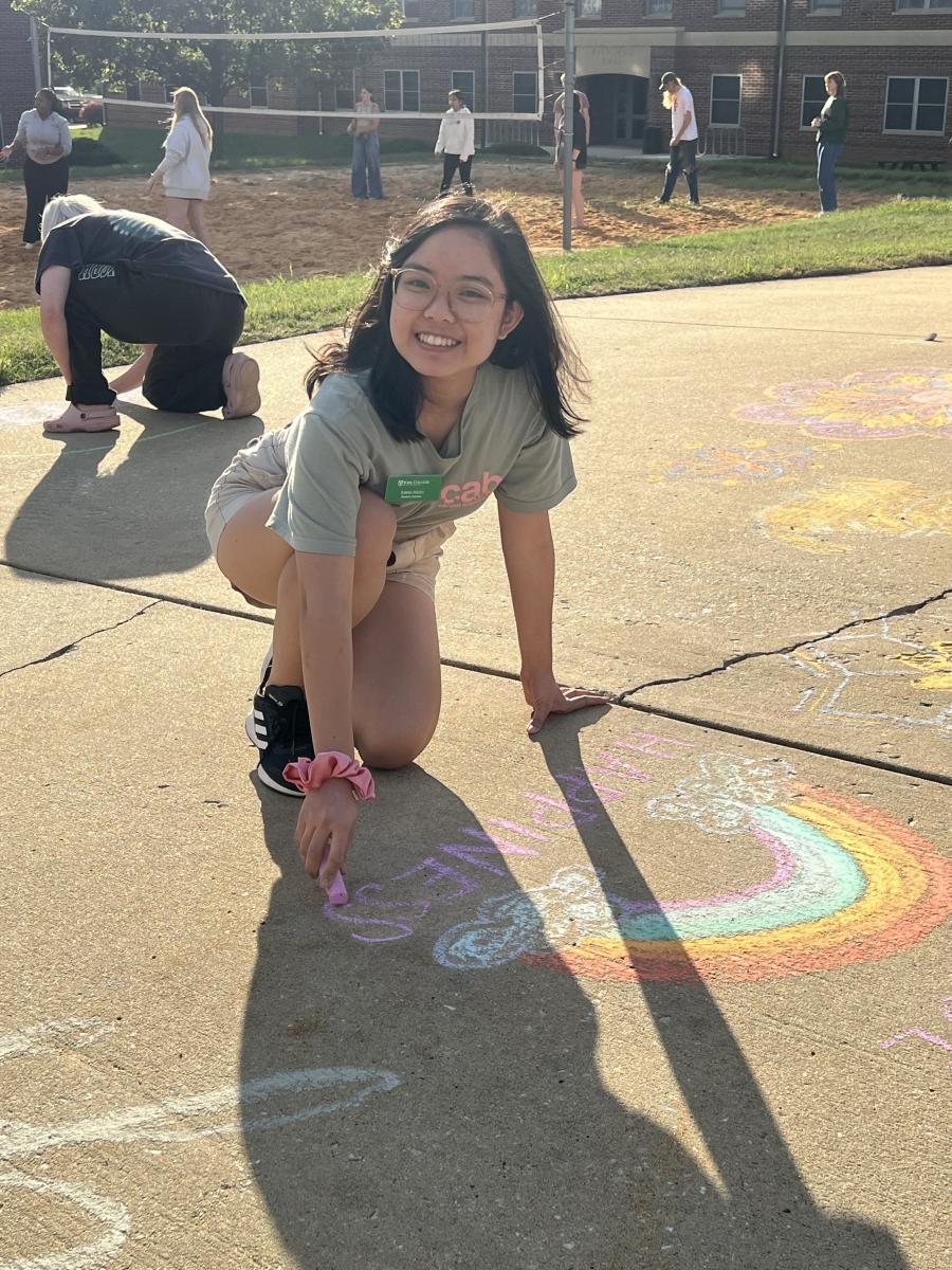 An individual drawing a rainbow with chalk on a sidewalk on a sunny day. People can be seen playing volleyball in the background.