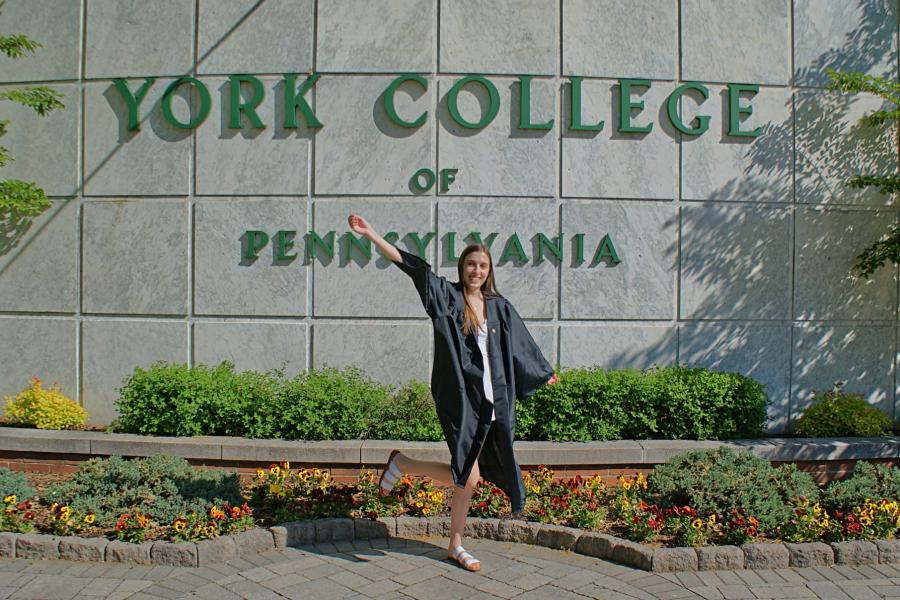 Student wearing a graduation gown poses joyfully in front of a "York College of Pennsylvania" sign.