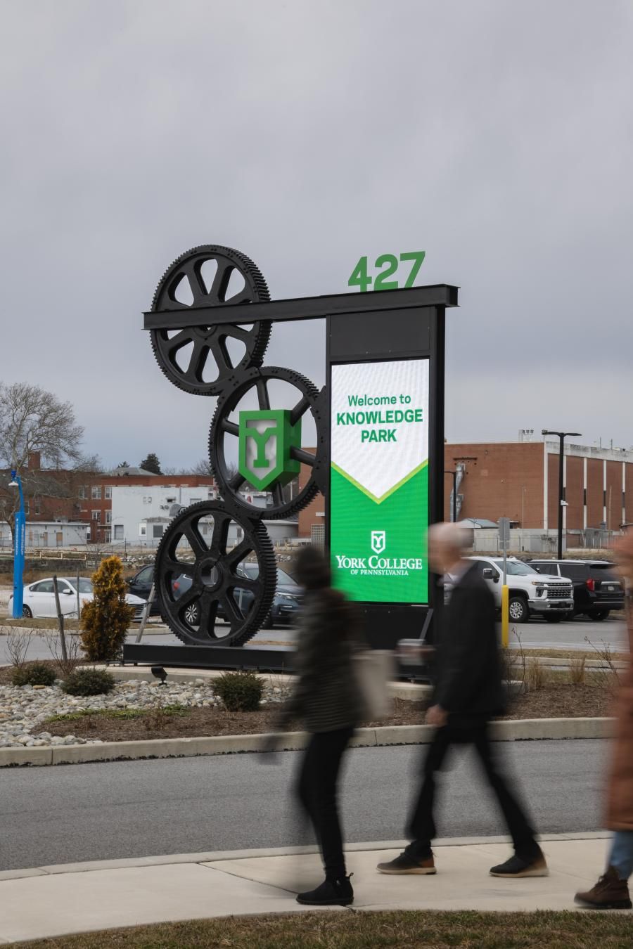A large, outdoor, metal sign with a digital display reading "Welcome to Knowledge Park." A few people can be seen walking on a sidewalk in the foreground.