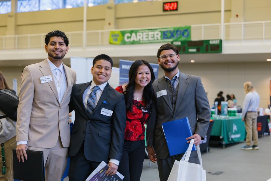 A group of college students in business attire smile widely at a professional networking event. A green banner reading "Career Expo" is visible in the background.
