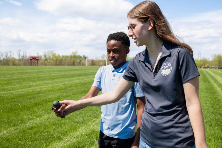 A college student wearing a grey polo shirt with a drone logo assists a younger child with a drone. They are outdoors on a sunny day.