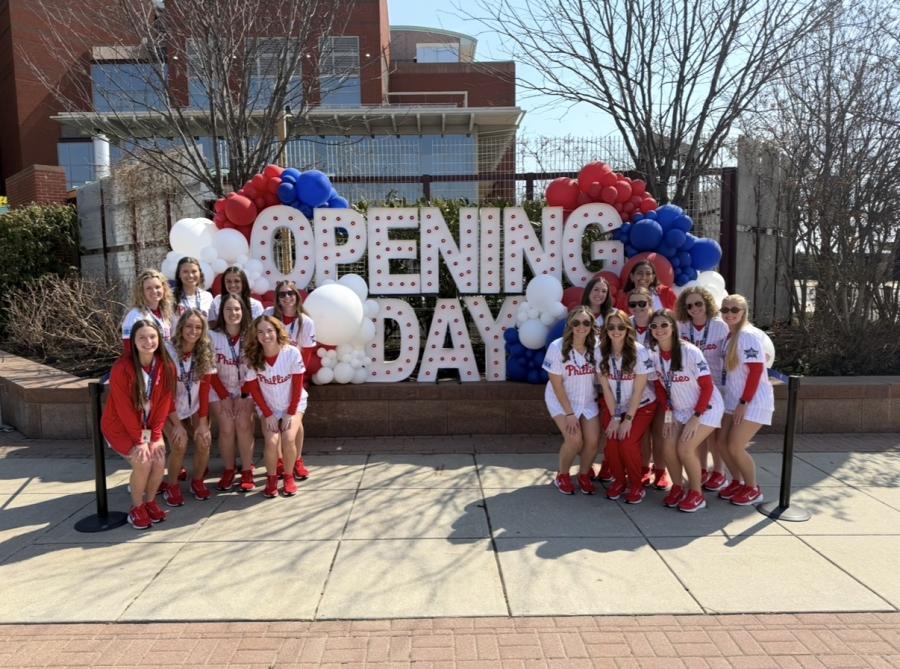 A group of people wearing Philadelphia Phillies uniforms posing in front of an "Opening Day" sign.