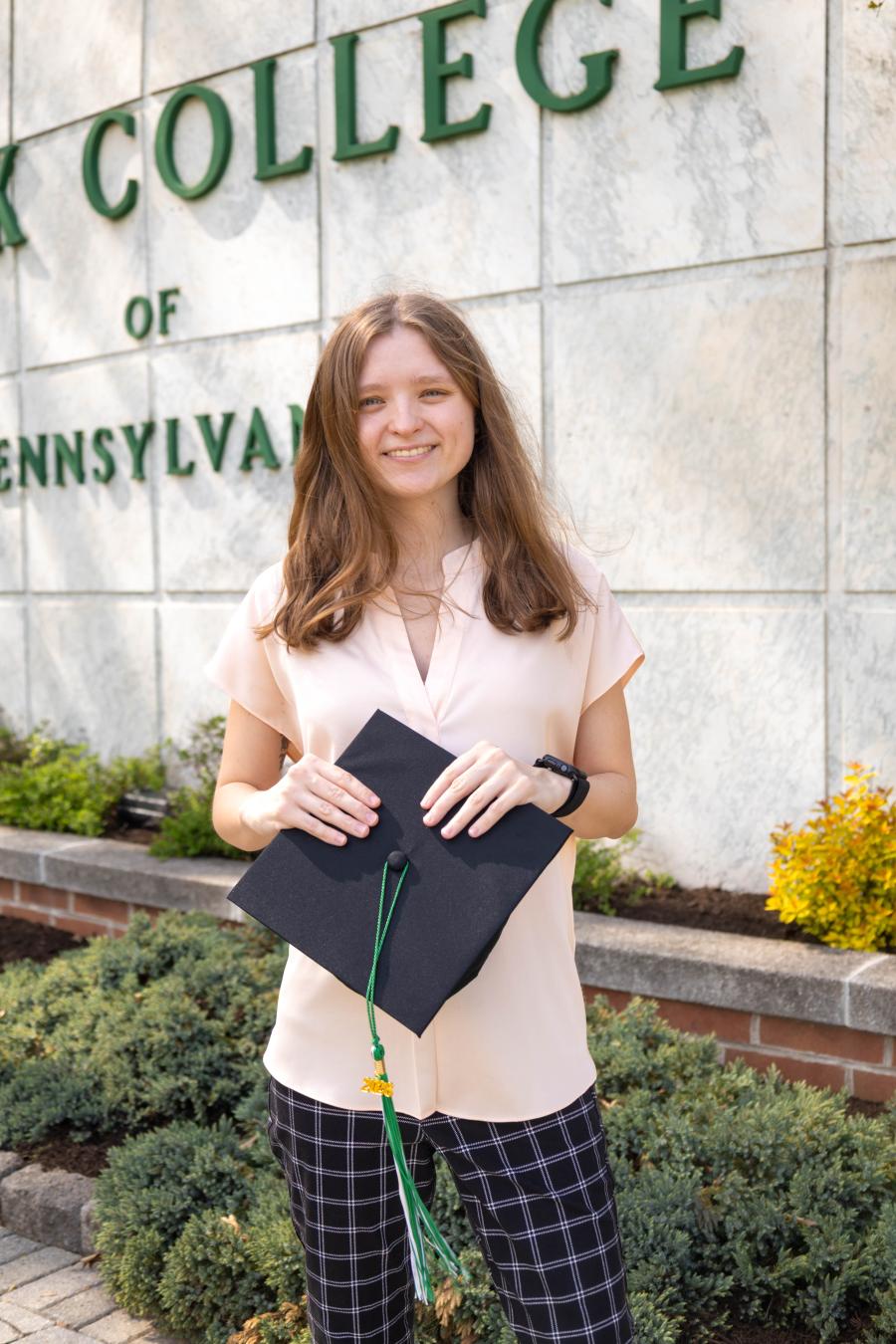 Student wearing a light colored top and dark, plaid pants smiles while holding a graduation cap. The York College of PA sign is partially visible in the background.