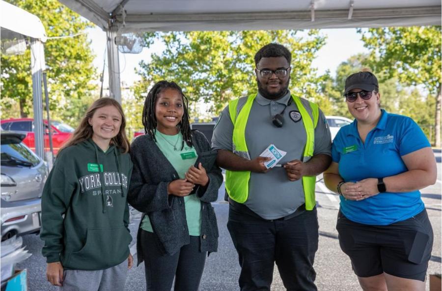 Four individuals smile at the camera beneath a canopy tent in a parking lot. They each have name tags and ID badges visible on their clothing.