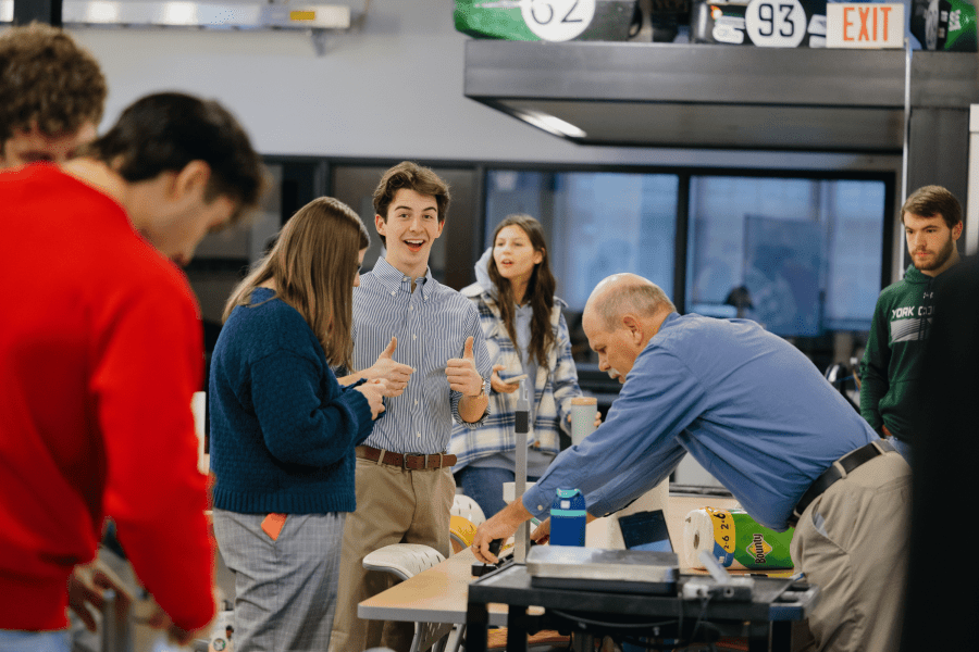 Aidan Heck ’27 gives two thumbs up while working alongside classmates and a faculty member during a Mechanical Engineering project presentation.