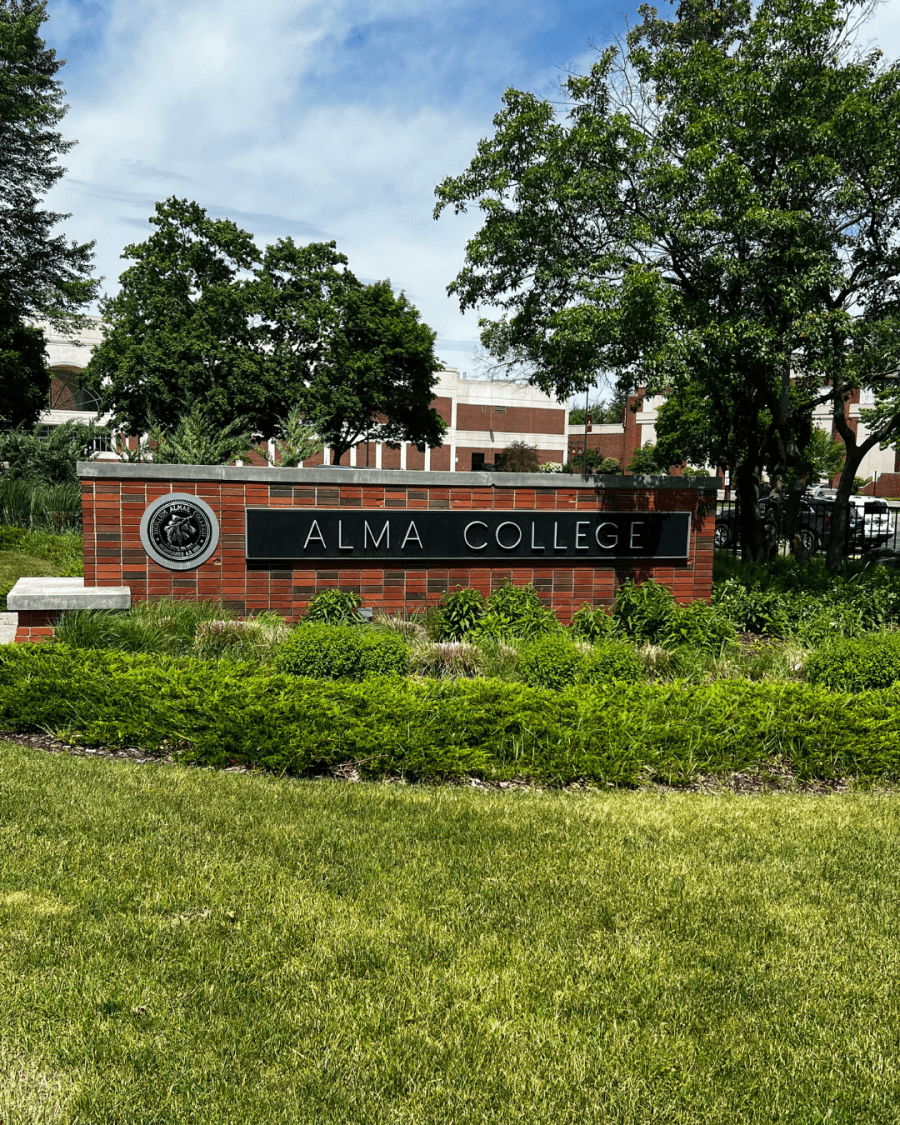 An Alma College campus sign is displayed in front of brick buildings, trees, and landscaping on a sunny day.