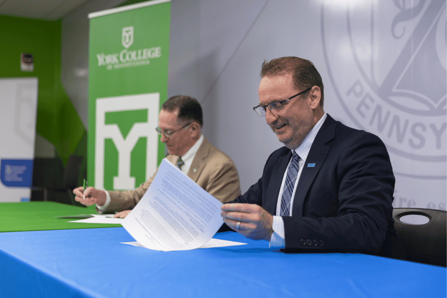 wo representatives review and sign documents at a blue-and-green table during a partnership signing event, with a York College of Pennsylvania banner and Pennsylvania graphic in the background.