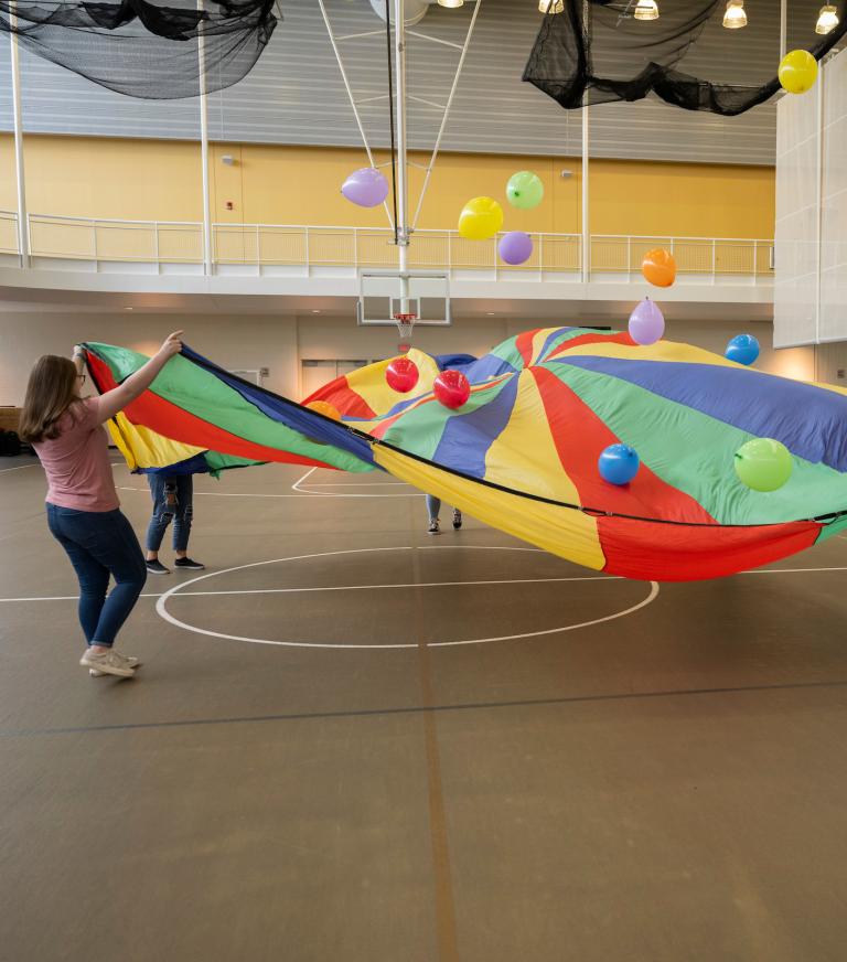 A group of students hold a large circular, multi-colored sheet bouncing balloons off of it.