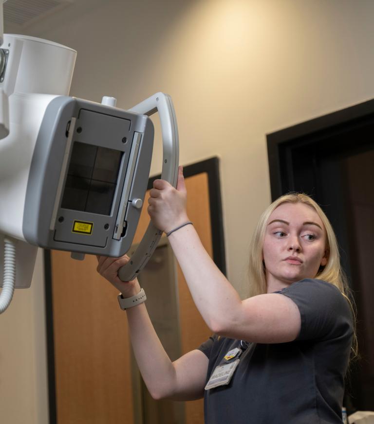 A student wearing scrubs adjusts a large overhead x-ray machine in a hospital setting.