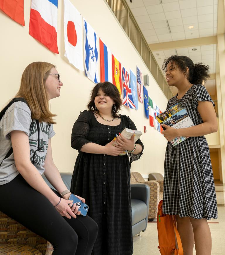 Three students talk in a hallway lined with flags from multiple countries.