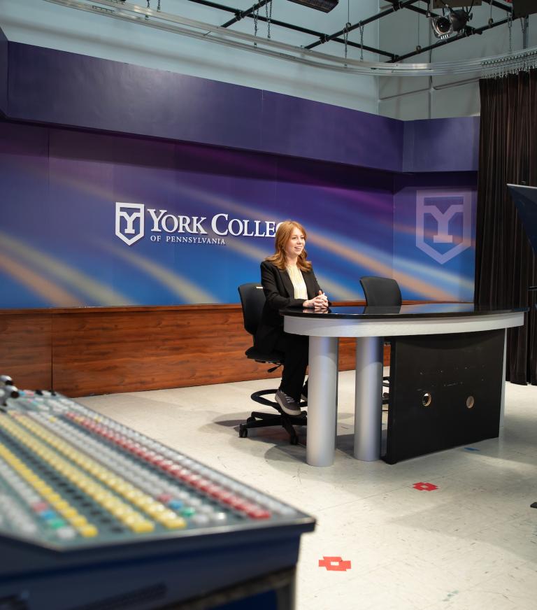 A student sits at a mock television news desk smiling and facing a video camera.