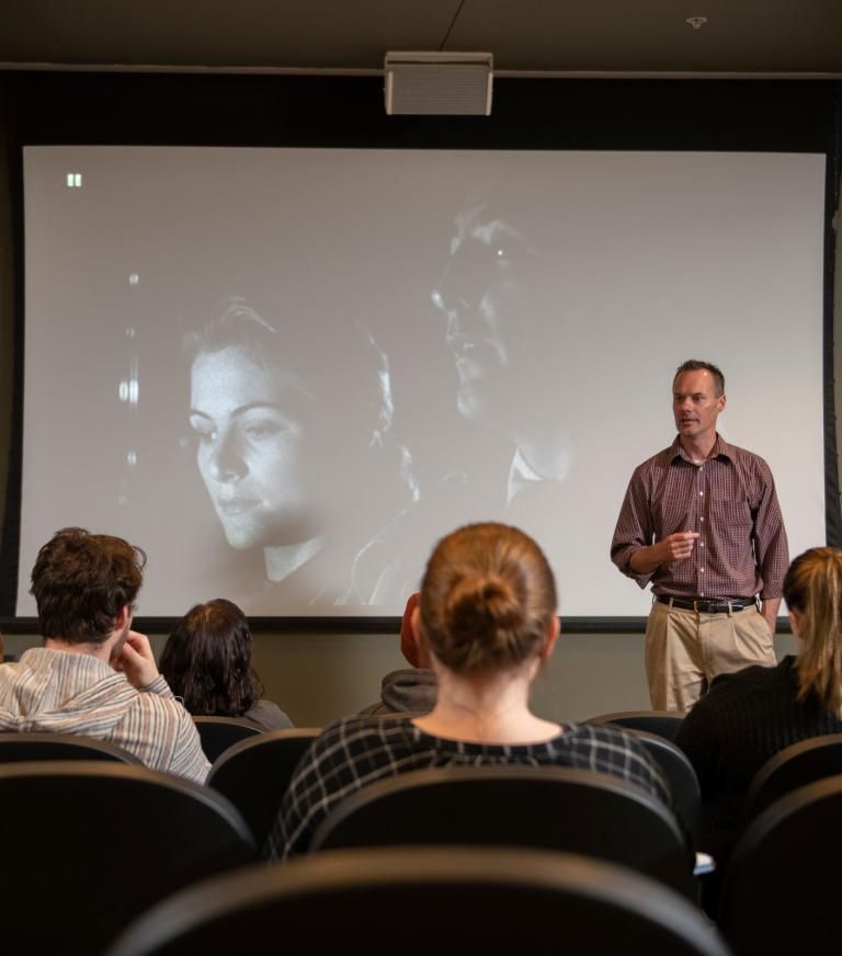 An instructor leads a class in front of a black and white film on a large screen