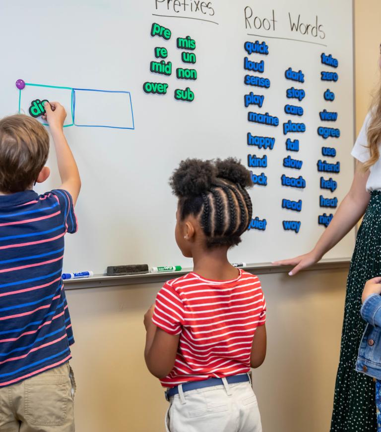 A teacher and three young children stand at a whiteboard. A child places magnetic label of a prefix in a diagram drawn on the whiteboard.