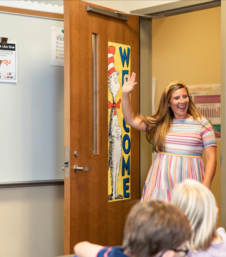 A teacher walking into a room waving at students.