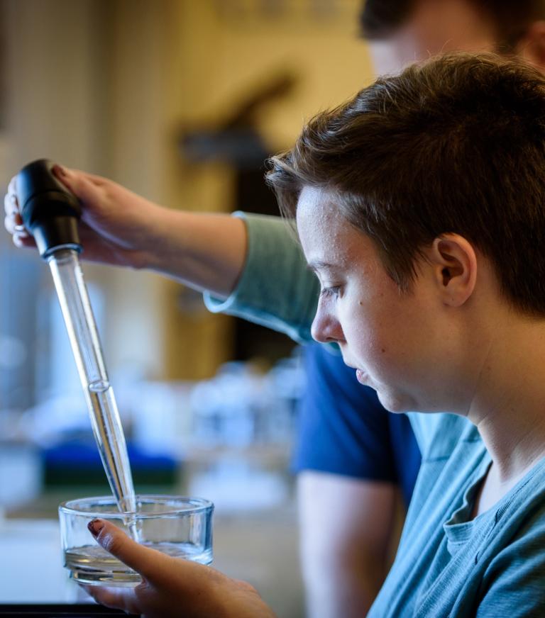 A student holds a large pipette in one hand in a glass container of liquid held in the other hand.