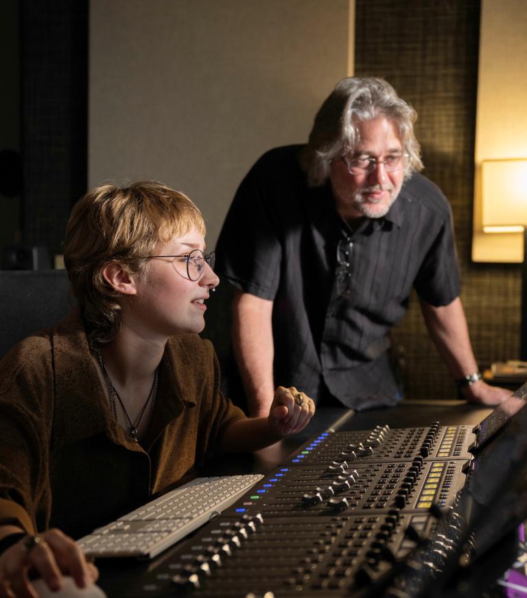 An instructor looks on as a student uses a mouse work on a music recording.