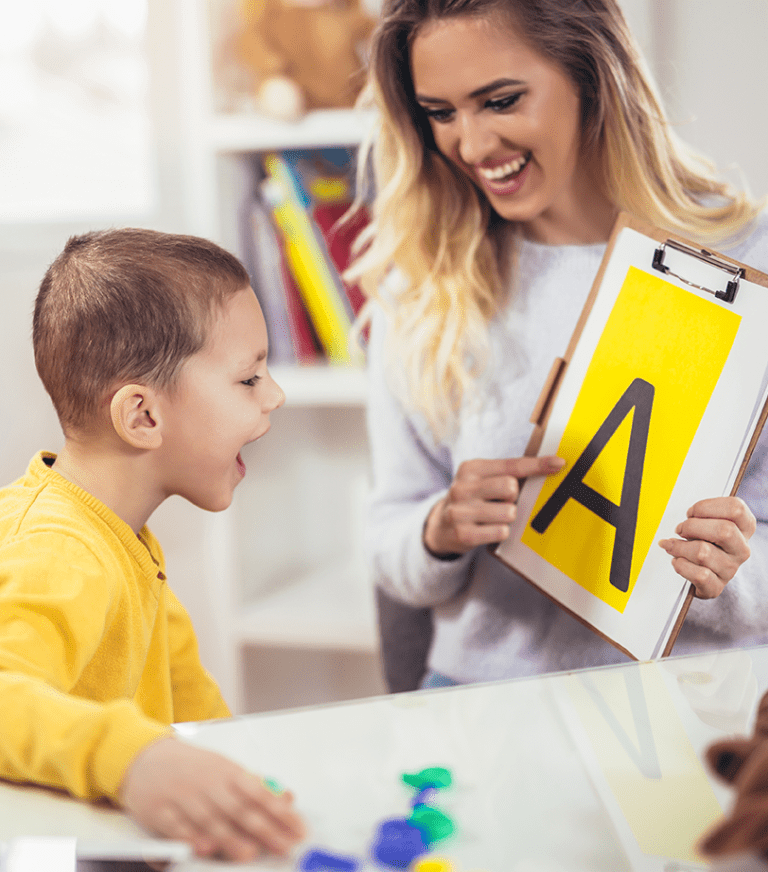 A teacher holds up a large printed letter A as a child in her classroom smiles.