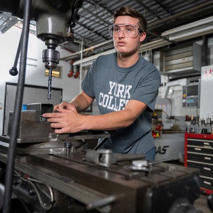 A student wearing safety glasses aligns a metal block on a drill press.