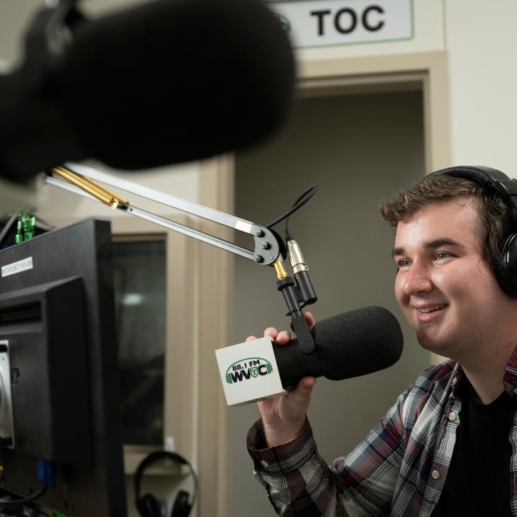 A student sits wearing headphones and holding a boom microphone facing a video camera.