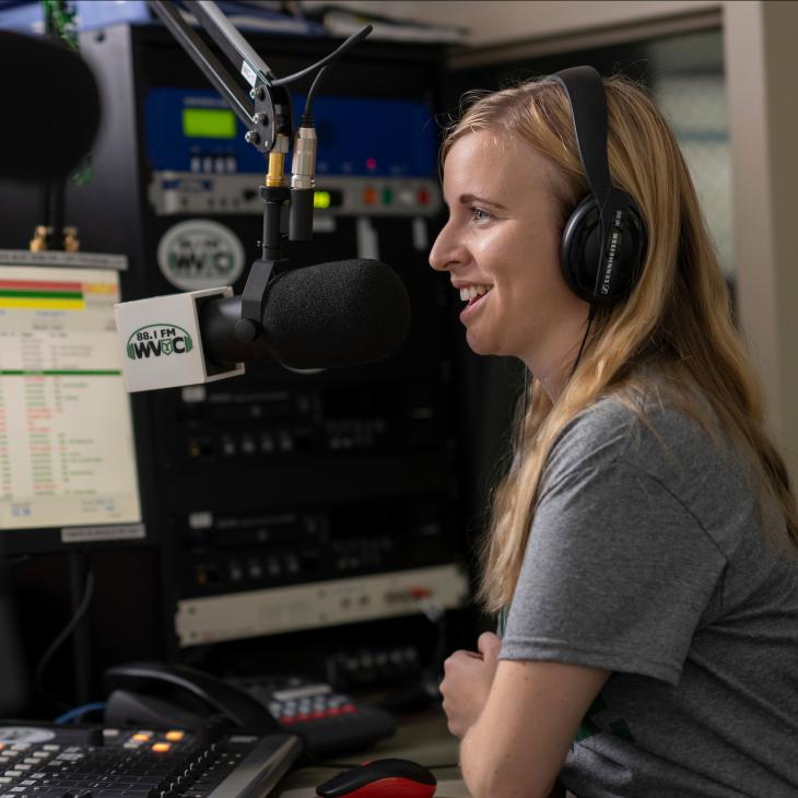A student wears over-ear headphones while sitting amid equipment in the campus radio station.