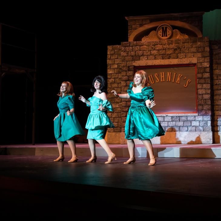 Three students sing on stage wearing 60's-style costume dresses during a performance of Little Shop of Horrors.