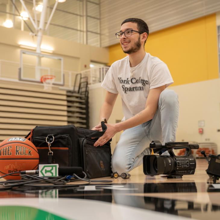 A student kneels by a camera equipment bag with a basketball and some camera equipment on the floor.