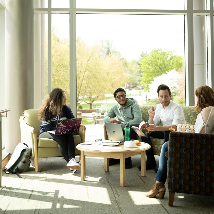 A group of three students and a professor sit in a lounge area on campus and chat, surrounded by floor-to-ceiling bay windows.
