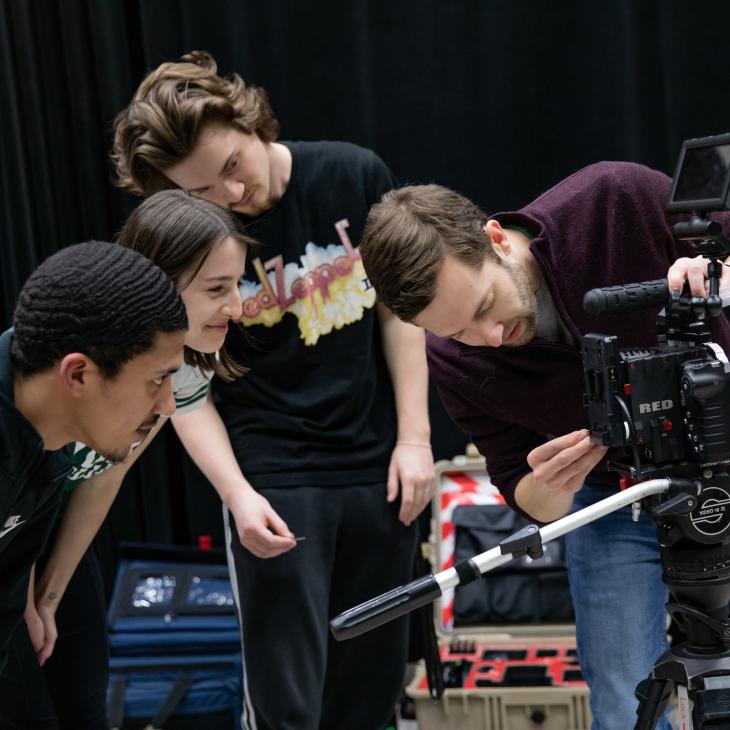 A group of students watch while a student sets up a video camera on a tripod.
