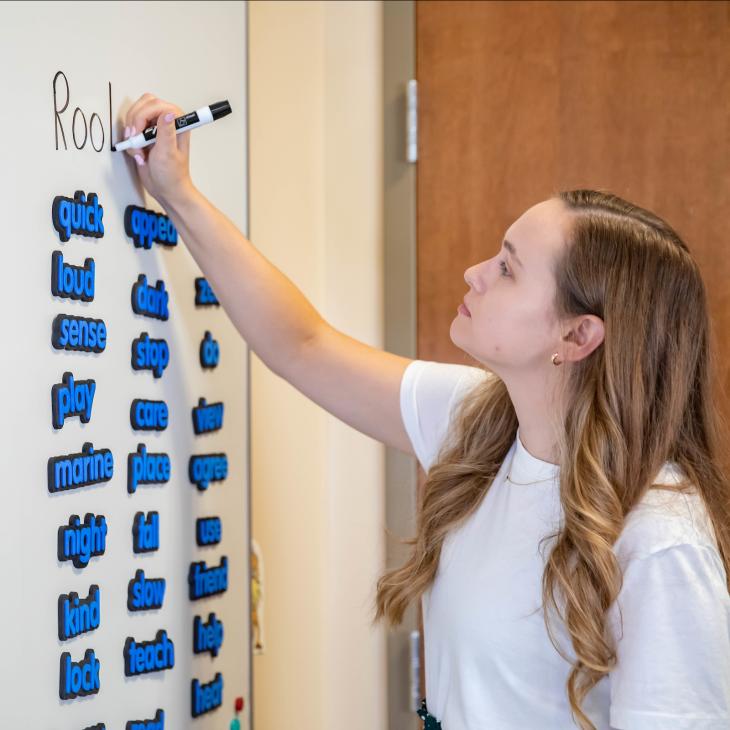 A teacher writes on a white board, writing the word "Roots" above a series of magnetic words and letters.