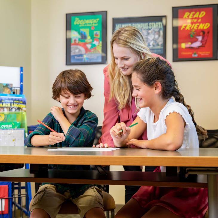 A teacher leans forward on a table behind two young students working on a project.
