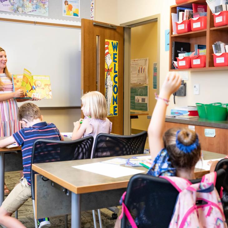 A teacher is standing in front of a classroom with 4 kids, two of the kids are raising their hands