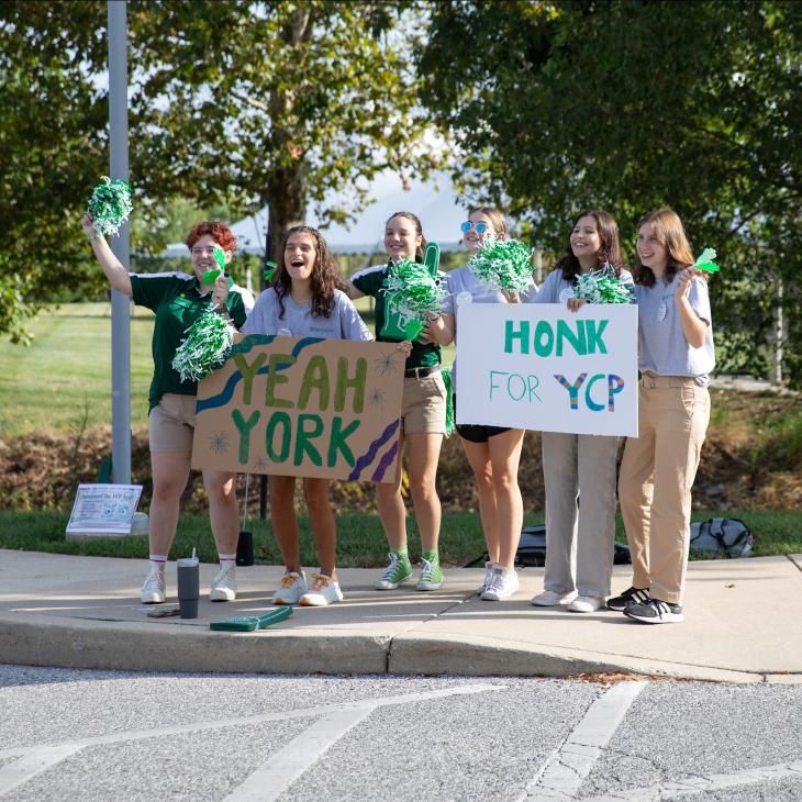 A group of student orientation leaders hold signs that say "Yeah York" and "Honk for YCP" as they greet new students driving into campus.
