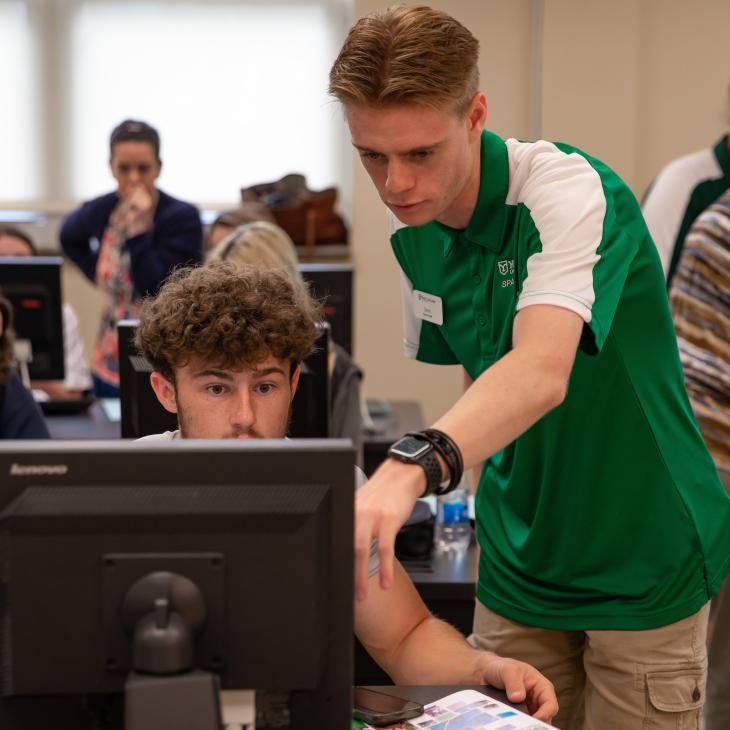 A student peer advisor points at a screen in the computer lab while assisting a new student.