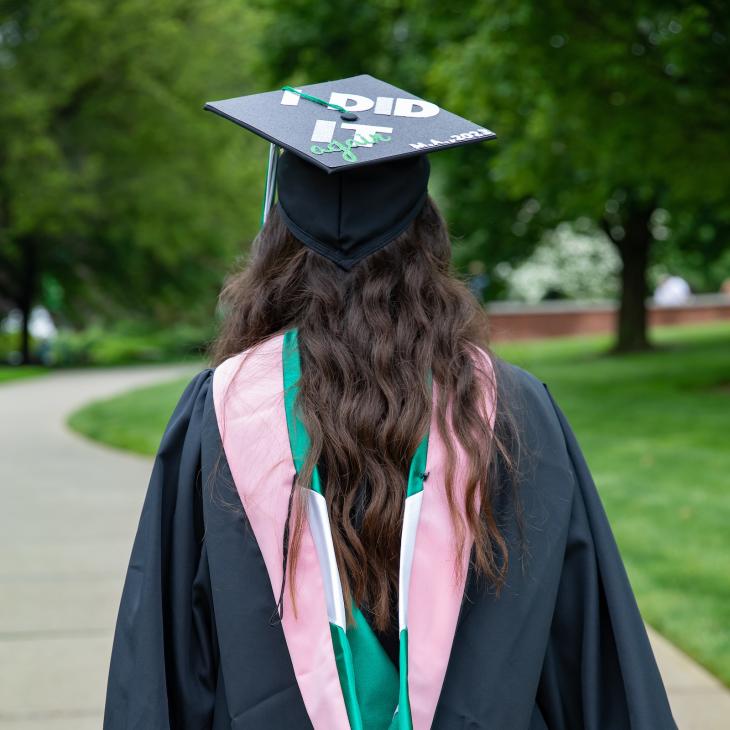 A student wearing graduation regalia walks away from the camera, down a campus sidewalk. Her cap decoration reads "I did it."