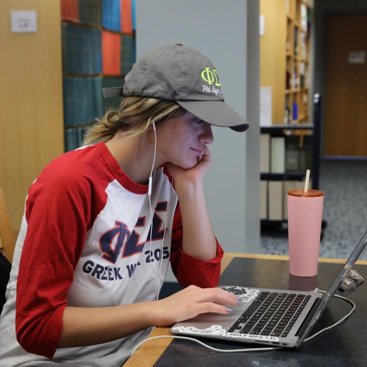 A student sits at a table in the library working on a laptop. The student is wearing a white sweatshirt with red sleeves and a green baseball cap. 