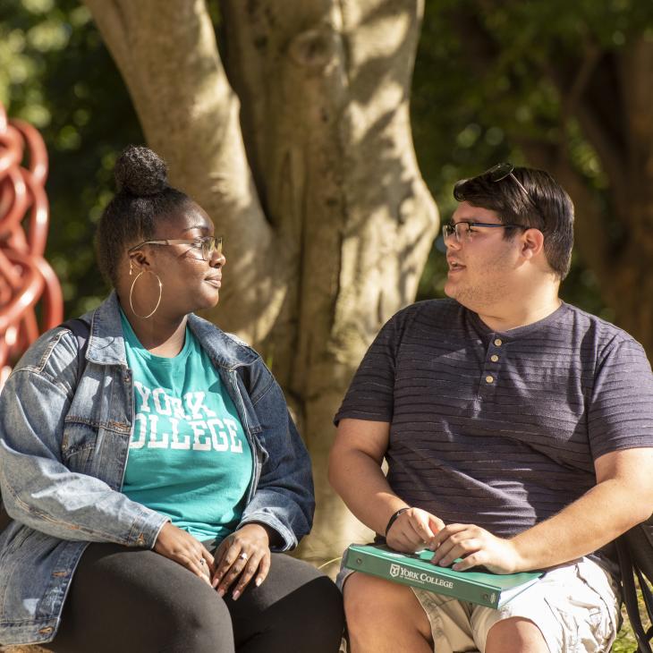 Two students converse sitting on a bench.