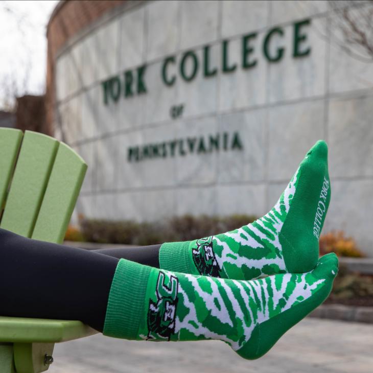 Feet are shown dangling off the edge of an Adirondack chair on campus, wearing a pair of green and white striped socks with a York College logo.