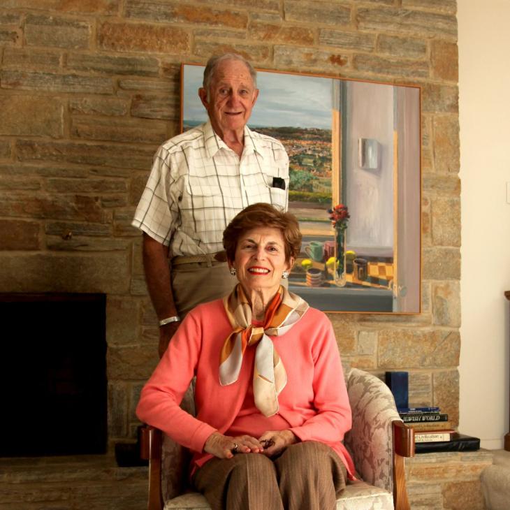 Doris and Bernard Gordon, the benefactors of the Doris and Bernard Gordon Center for Jewish Student Life. Doris is seated wearing a salmon colored sweater while Bernard stands behind the chair in white windowpane pattern shirt.